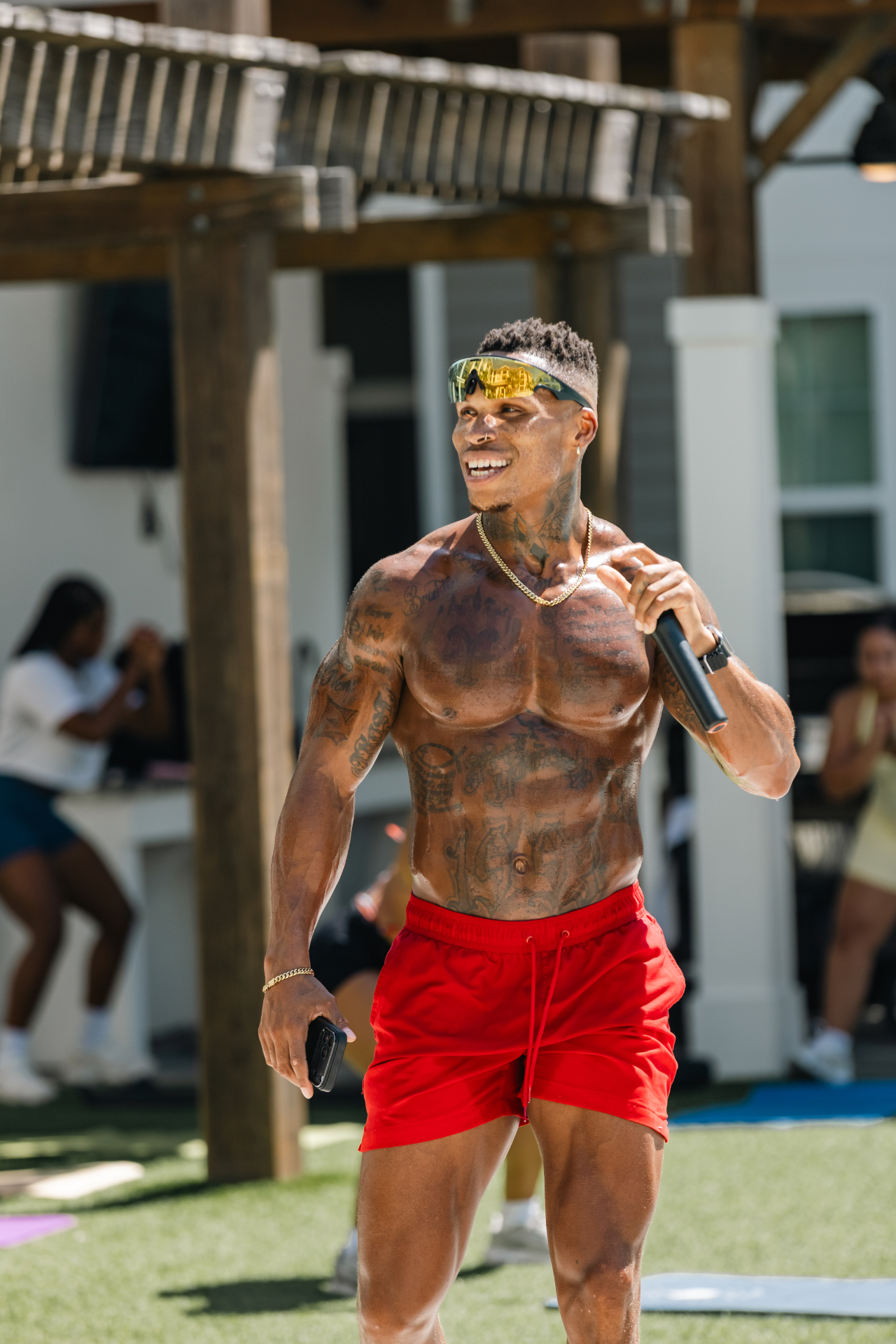 Coach Mac smiling with a microphone in red shorts at an outdoor training event
