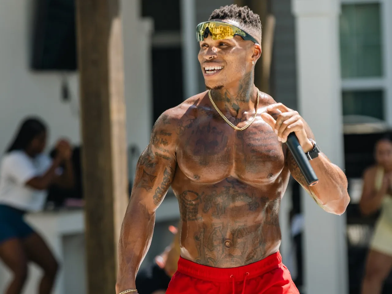 Coach Mac smiling with a microphone in red shorts at an outdoor training event